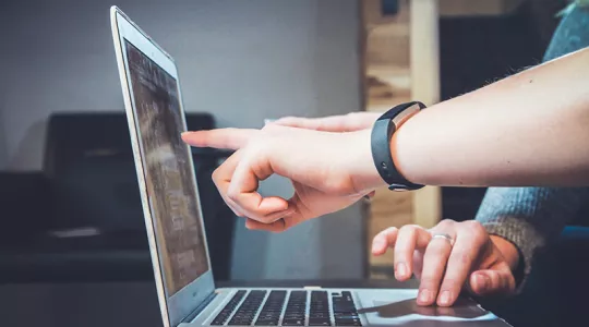 White walls, laptop on a desk in the background, two people at a desk; one of them pointing at a laptop screen facing right and the other's hand on the keyboard
