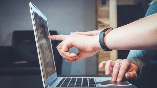 White walls, laptop on a desk in the background, two people at a desk; one of them pointing at a laptop screen facing right and the other's hand on the keyboard