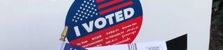 Sign on white cardboard with the "I Voted" circle logo in the middle, a USA flag, a "I Voted" sticker, and a ballot envelope held under it