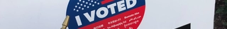 Sign on white cardboard with the "I Voted" circle logo in the middle, a USA flag, a "I Voted" sticker, and a ballot envelope held under it